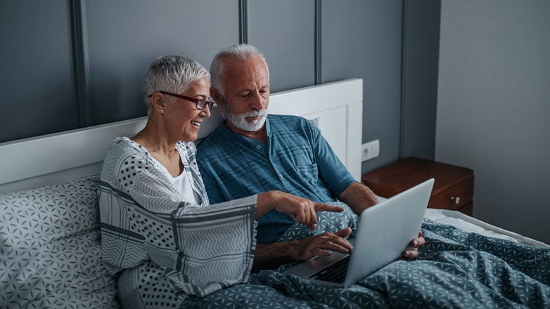 old couple sitting in bed pointing and working on laptop