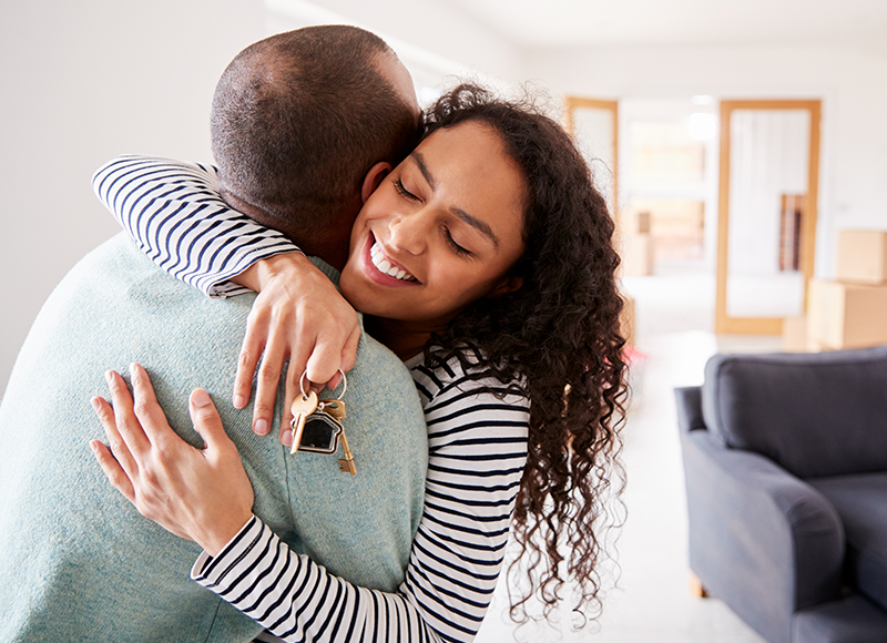 A young couple is hugging each other. The woman is holding a set of keys in her hand. They are standing in a living room that is mostly empty, except for a couch and a few boxes.