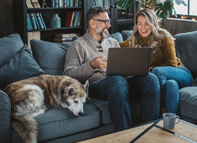 Mature couple looks at a laptop while relaxing on their couch with their pet dog.